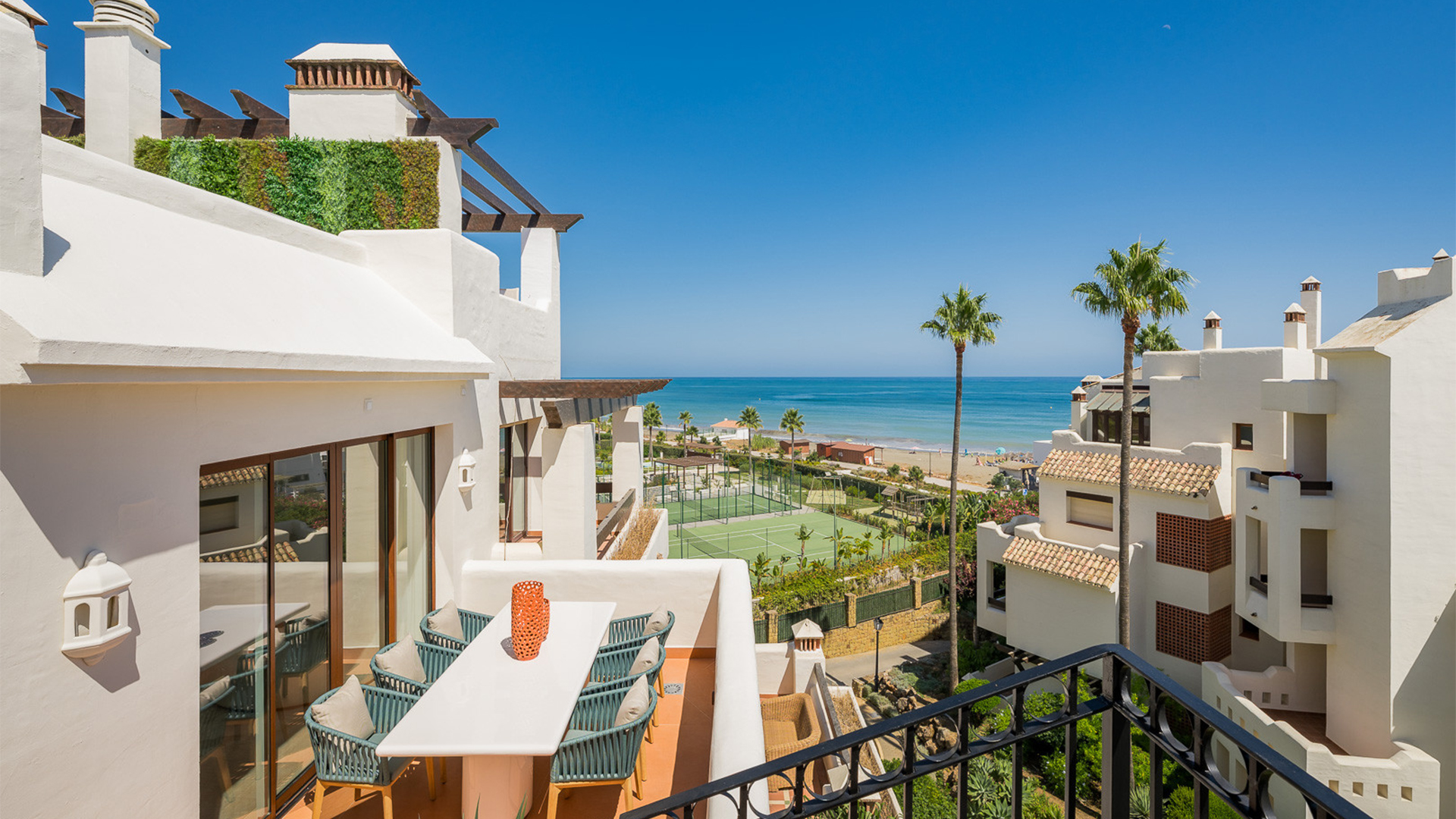 Luxury beachfront penthouse in Estepona with private balcony, outdoor dining area, tennis court views, palm trees, and stunning Mediterranean Sea outlook